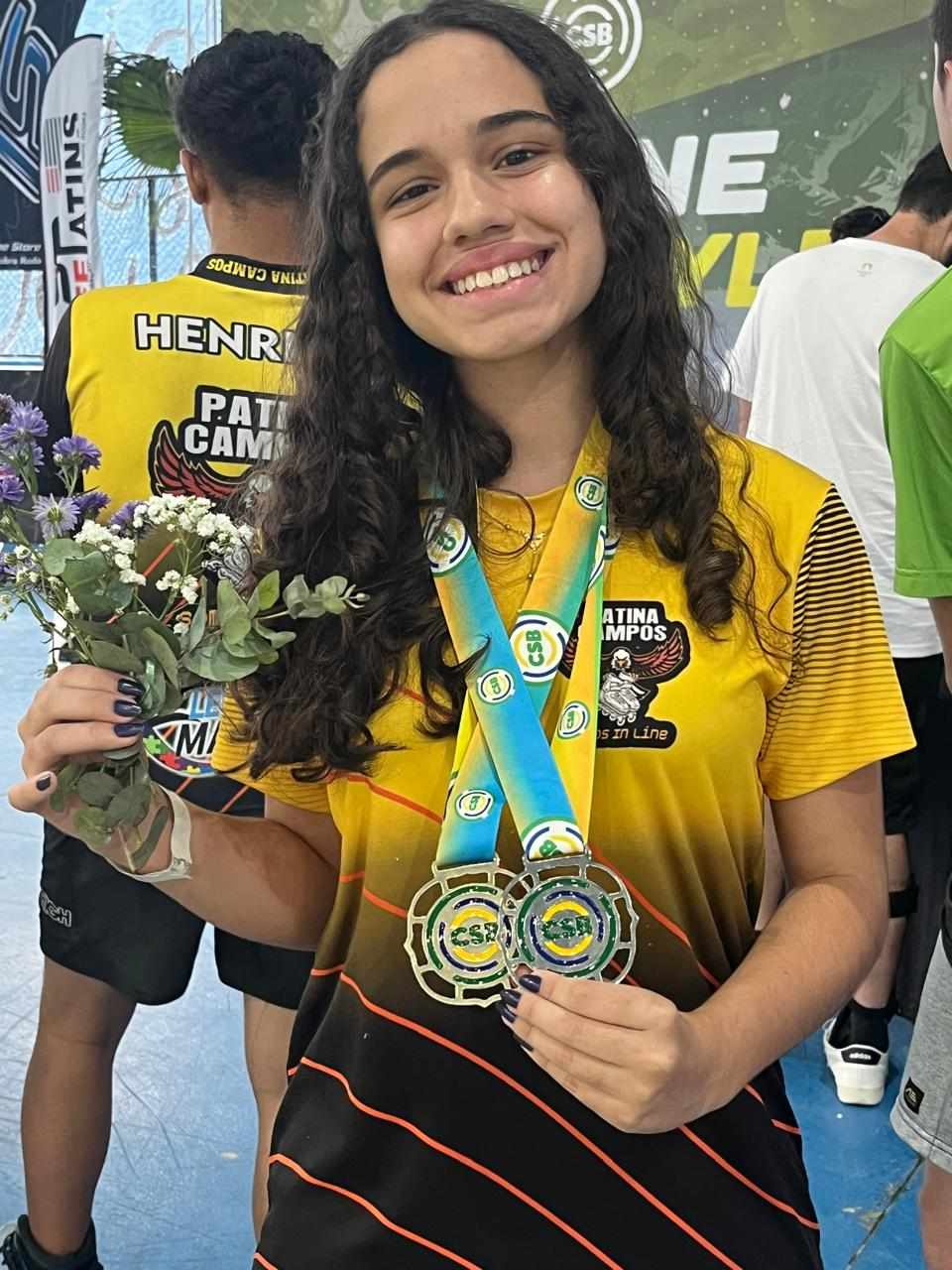 Yasmin sorrindo com duas medalhas de ouro no peito e um buquê de flores